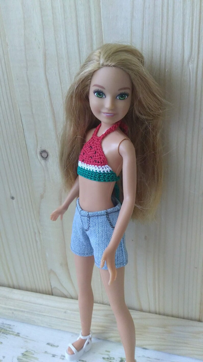 May include: A doll with long blonde hair wearing a red, green and white watermelon-patterned halter top, blue denim shorts and white sandals.