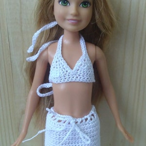 May include: White crocheted bikini top and skirt set for a doll. The top has a halter style and the skirt is a mini skirt with a tie at the waist.