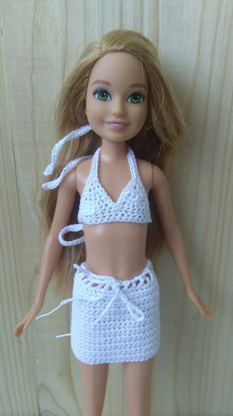 May include: White crocheted bikini top and skirt set for a doll. The top has a halter style and the skirt is a mini skirt with a tie at the waist.