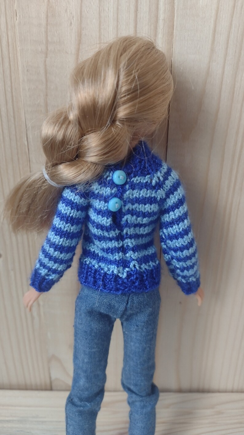 May include: A blue and light blue striped knitted sweater with blue buttons. The sweater is being worn by a doll with blonde hair.