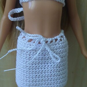 May include: White crocheted bikini top and skirt set for a doll. The top is halter style and the skirt is a wrap style with a tie at the waist.