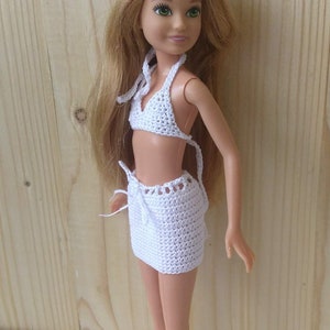 May include: A white crocheted bikini top and skirt set for a doll. The doll has long blonde hair and green eyes.