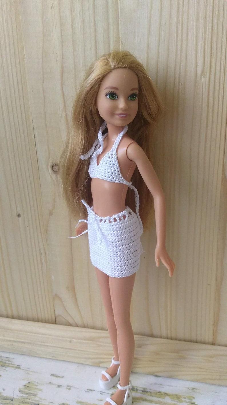 May include: A white crocheted bikini top and skirt set for a doll. The doll has long blonde hair and green eyes.