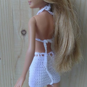 May include: A white crocheted bikini top and skirt set for a doll. The top has a halter style and the skirt is a mini skirt. The doll has long blonde hair and green eyes.