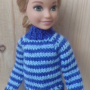 May include: A doll wearing a blue and light blue striped knitted sweater with a turtleneck. The doll is wearing blue jeans.