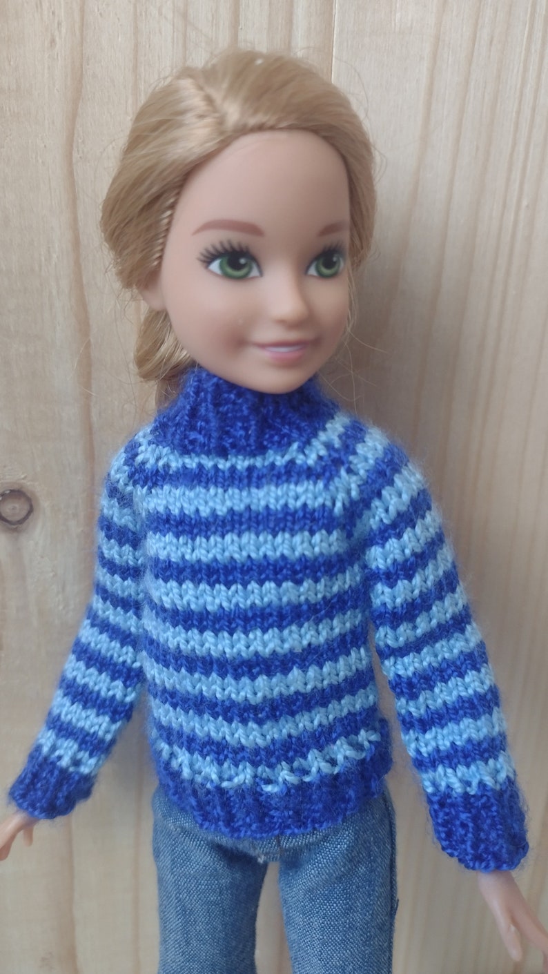 May include: A doll wearing a blue and light blue striped knitted sweater with a turtleneck. The doll is wearing blue jeans.