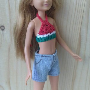May include: A blonde doll wearing a red, white and green crocheted watermelon top and blue denim shorts.