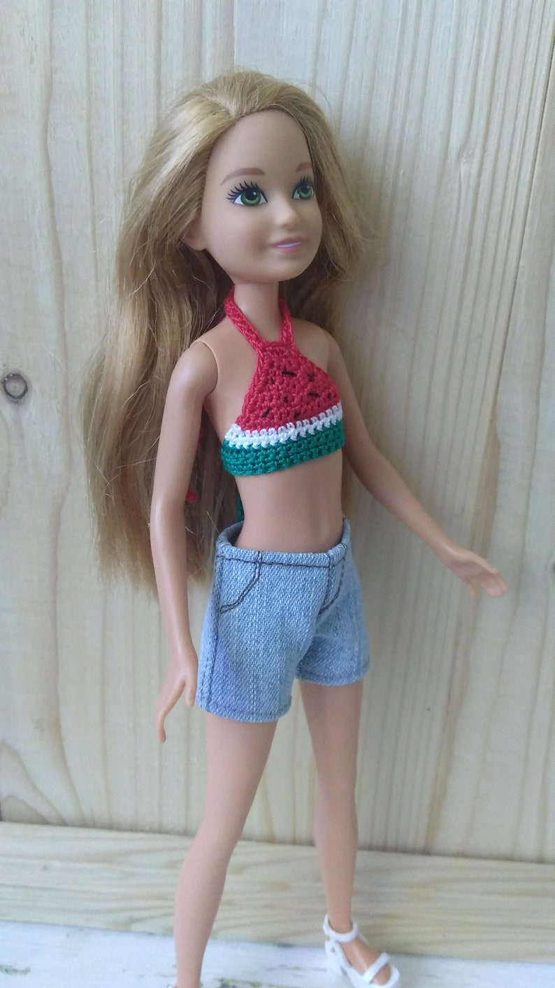 May include: A blonde doll wearing a red, white and green crocheted watermelon top and blue denim shorts.