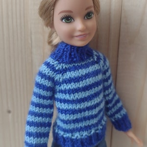 May include: A doll wearing a blue and light blue striped knitted sweater with a turtleneck and blue jeans.
