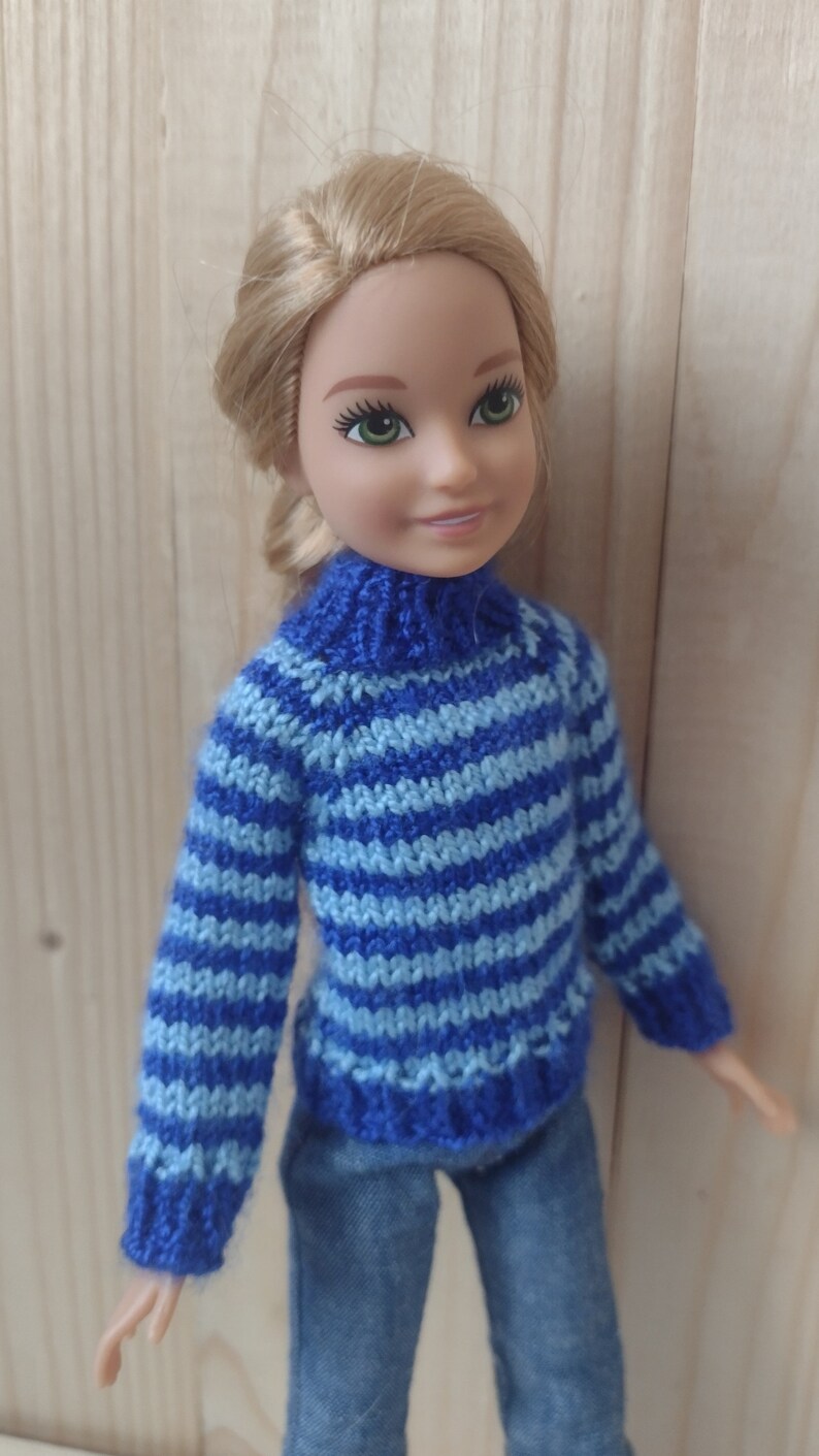 May include: A doll wearing a blue and light blue striped knitted sweater with a turtleneck and blue jeans.
