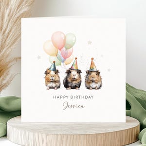 May include: A square greeting card featuring a watercolour illustration of three guinea pigs wearing colourful party hats. Above them are pastel-coloured balloons and small stars. The text reads HAPPY BIRTHDAY and the name Jessica in script font.