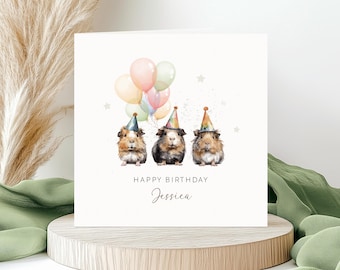 Puede incluir: Una tarjeta de felicitación cuadrada con una ilustración en acuarela de tres cobayas con gorros de fiesta coloridos. Encima hay globos de tonos pastel y pequeñas estrellas. El texto dice HAPPY BIRTHDAY y el nombre Jessica en cursiva.