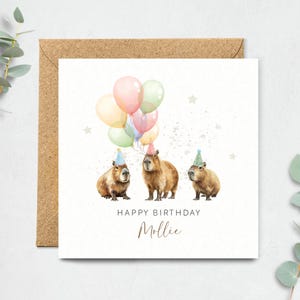 May include: A birthday card featuring three capybaras wearing party hats and holding colorful balloons. The words "Happy Birthday Mollie" are written below the capybaras in a handwritten font. The card is on a white background with a brown envelope.