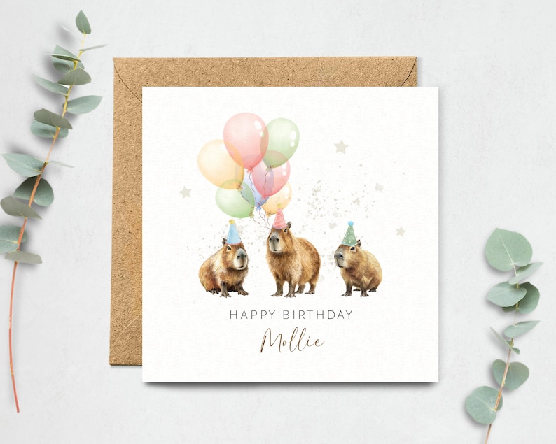May include: A birthday card featuring three capybaras wearing party hats and holding colorful balloons. The words "Happy Birthday Mollie" are written below the capybaras in a handwritten font. The card is on a white background with a brown envelope.