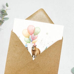 May include: A birthday card featuring a capybara wearing a party hat and holding balloons in pastel colors. The card is inside a brown envelope, with eucalyptus leaves in the background.