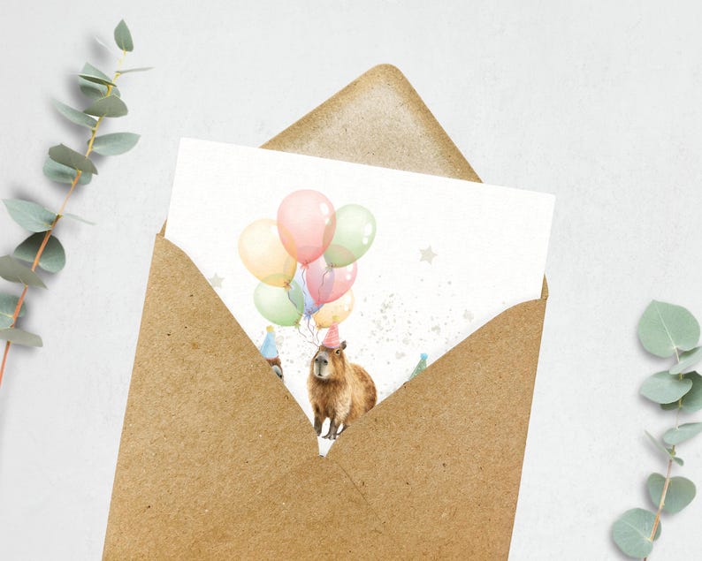 May include: A birthday card featuring a capybara wearing a party hat and holding balloons in pastel colors. The card is inside a brown envelope, with eucalyptus leaves in the background.