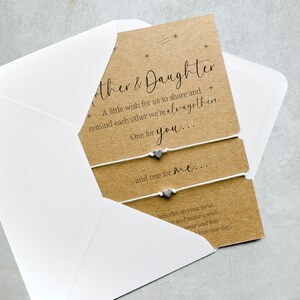 May include: A brown card with white string bracelets and silver heart charms. The card has the text "Mother & Daughter" and a message about sharing a wish and reminding each other that they are always there.