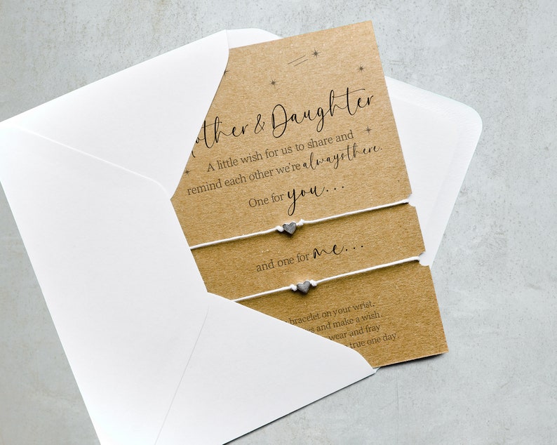 May include: A brown card with white string bracelets and silver heart charms. The card has the text "Mother & Daughter" and a message about sharing a wish and reminding each other that they are always there.