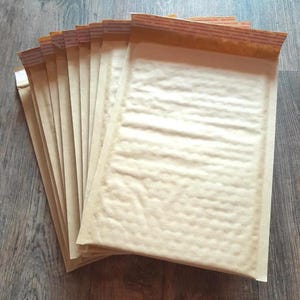 May include: A stack of rectangular tan paper bubble mailers arranged in a fan pattern. Each mailer features a textured surface with a protective interior lining and a brown adhesive strip along the top edge for sealing the envelope securely.