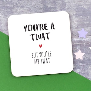 May include: A white square coaster with the text "YOU'RE A TWAT" above a red heart and the text "BUT YOU'RE MY TWAT" in black. The coaster is on a green surface with star-shaped confetti.
