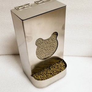 May include: A stainless steel pet food dispenser with a bear-shaped opening and a removable tray. The dispenser has a hinged lid and is filled with green pet food pellets.