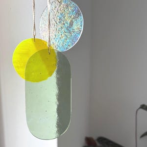 Könnte beinhalten: Ein hängendes Mobile mit drei verschiedenen Glasformen, die an weißen Schnüren befestigt sind. Die Elemente bestehen aus einer gelben Glasscheibe, einer schillernden, strukturierten Glasscheibe und einem glatten, länglichen ovalen Stück in blassem Salbeigrün.