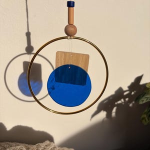 Peut inclure: Mobile moderne avec un anneau en métal doré, des perles en bois et des formes géométriques. Le design comprend un cercle bleu et un rectangle en bois clair. Un petit rectangle bleu est en haut. Le mobile est suspendu à une ficelle.