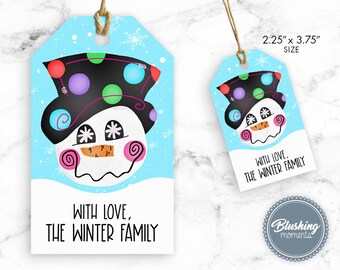 Puede incluir: Una etiqueta de regalo azul con una ilustración de muñeco de nieve. El muñeco de nieve lleva un sombrero negro con lunares de colores, tiene una cara blanca con una nariz de zanahoria naranja y mejillas rosadas. El texto en la etiqueta dice "Con amor, La familia de invierno".