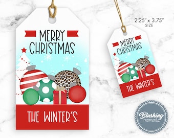 Puede incluir: Una etiqueta de regalo de Navidad roja y blanca con fondo azul y una estrella blanca. La etiqueta presenta un grupo de adornos navideños, incluyendo un adorno de rayas rojas y blancas, un adorno de lunares verdes y blancos, un adorno de estampado de leopardo y un adorno rojo. La etiqueta también incluye el texto "Feliz Navidad" y "The Winter's". La etiqueta mide 5,7 cm por 9,5 cm.
