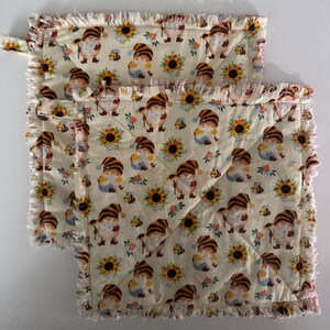 May include: Two quilted pot holders with a cream-colored background. The fabric is patterned with gnomes, sunflowers, and bees. The edges are trimmed with a fringed border. One pot holder has a small fabric loop for hanging.
