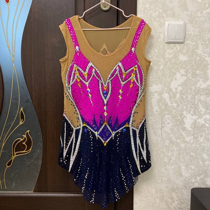 May include: A pink, blue, and gold leotard with a geometric design and rhinestones. The leotard has a nude mesh overlay and a scoop neck.