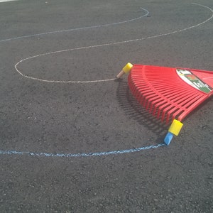 May include: A red plastic rake with a wooden handle is used to draw a blue chalk line on a black asphalt surface. The rake has yellow plastic handles on the end of the tines.