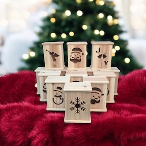 May include: A collection of small, wooden boxes with festive Christmas designs. Each box features a different laser-cut image, including a snowman, snowflake, Santa Claus, and an angel. The boxes are arranged on a plush, red surface, with a blurred Christmas tree in the background.