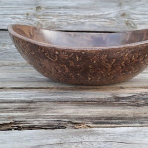 Op de afbeelding: Een handgemaakte, ovaalvormige bruine keramische kom met een getextureerde buitenkant. De kom heeft een gladde, glanzende binnenkant en staat op een verweerd houten oppervlak. Deze decoratieve kom is geschikt voor het serveren van voedsel of als decoratie.