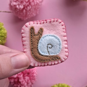 May include: A handmade felt snail patch. The patch is square with a pink background and a brown snail with a light blue shell. The snail is stitched with brown thread. The patch is surrounded by a pink stitched border. The background is pink.
