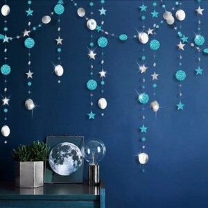 May include: Decorative garland with hanging silver, teal, and glittery blue shapes, including stars, circles, and moons, against a dark blue wall. A table displays a framed moon print, a potted plant, and a decorative light.