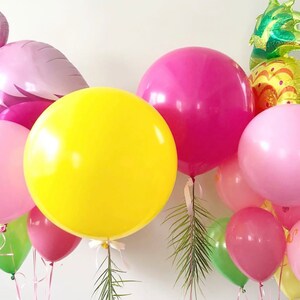 May include: A collection of colorful balloons, including a large yellow sphere, pink and magenta round balloons, and a flamingo-shaped balloon. A pineapple-shaped balloon is also visible, along with green leaves. Ribbons and strings are attached to the balloons.