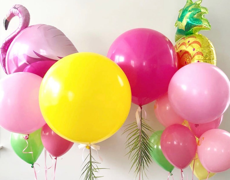 May include: A collection of colorful balloons, including a large yellow sphere, pink and magenta round balloons, and a flamingo-shaped balloon. A pineapple-shaped balloon is also visible, along with green leaves. Ribbons and strings are attached to the balloons.