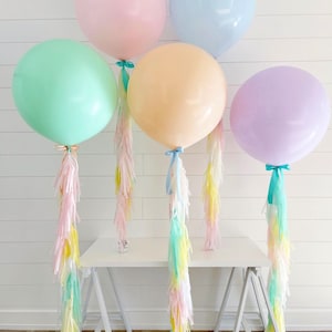 May include: Five large pastel-colored balloons: pink, blue, green, peach, and purple. Each balloon is tied with a ribbon and has a long tassel of pastel-colored streamers. The balloons are arranged on a white table against a white background.