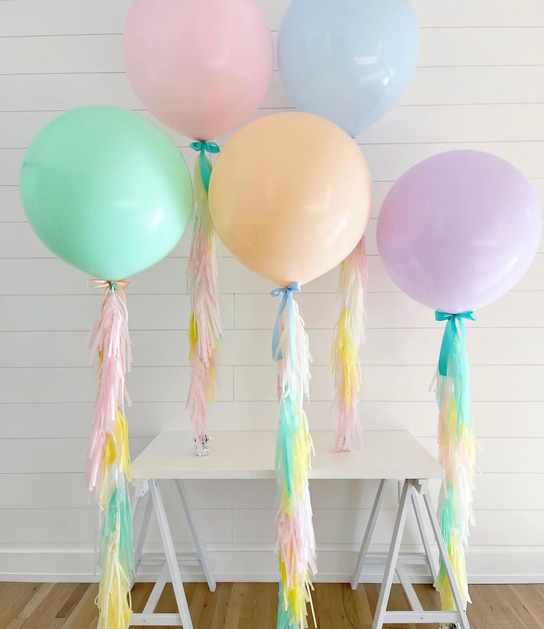 May include: Five large pastel-colored balloons: pink, blue, green, peach, and purple. Each balloon is tied with a ribbon and has a long tassel of pastel-colored streamers. The balloons are arranged on a white table against a white background.