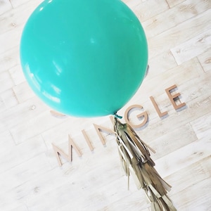 May include: A large, round, teal balloon with a gold tassel tail. The word "MING LE" is spelled out in wooden letters on a white wood-planked surface. Party decoration.
