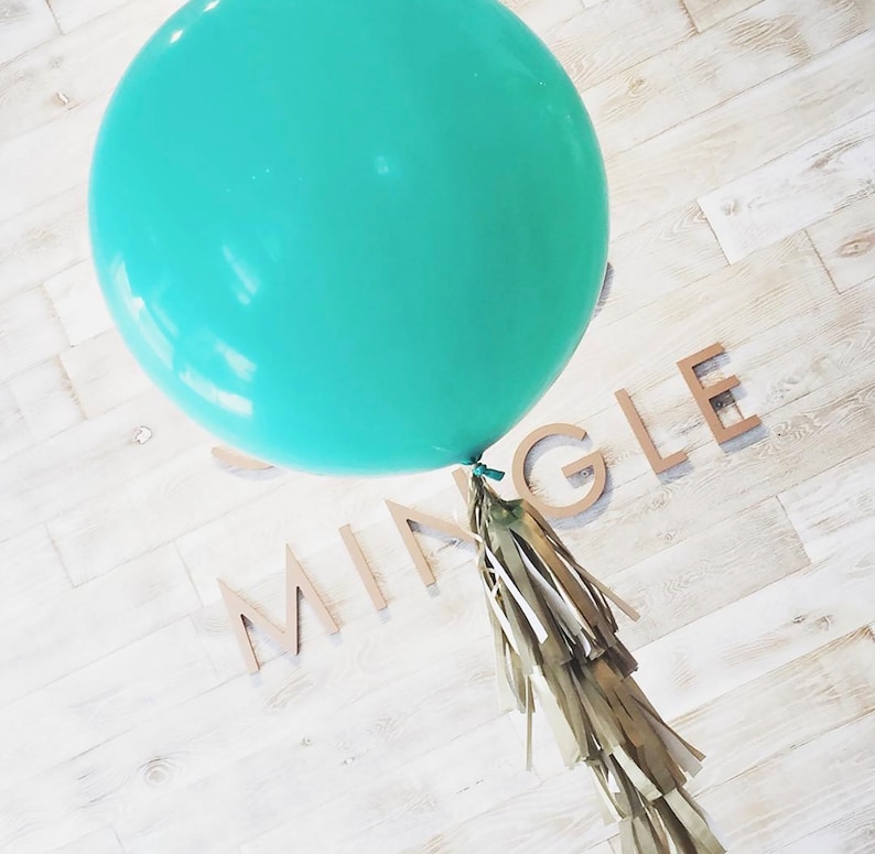 May include: A large, round, teal balloon with a gold tassel tail. The word "MING LE" is spelled out in wooden letters on a white wood-planked surface. Party decoration.