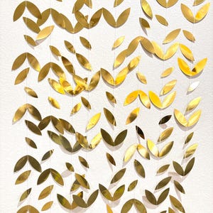 May include: A decorative wall hanging featuring numerous gold-colored leaf-shaped cutouts. The leaves are suspended from thin strings, creating a layered effect. The leaves have a metallic sheen and are arranged in a cascading pattern.