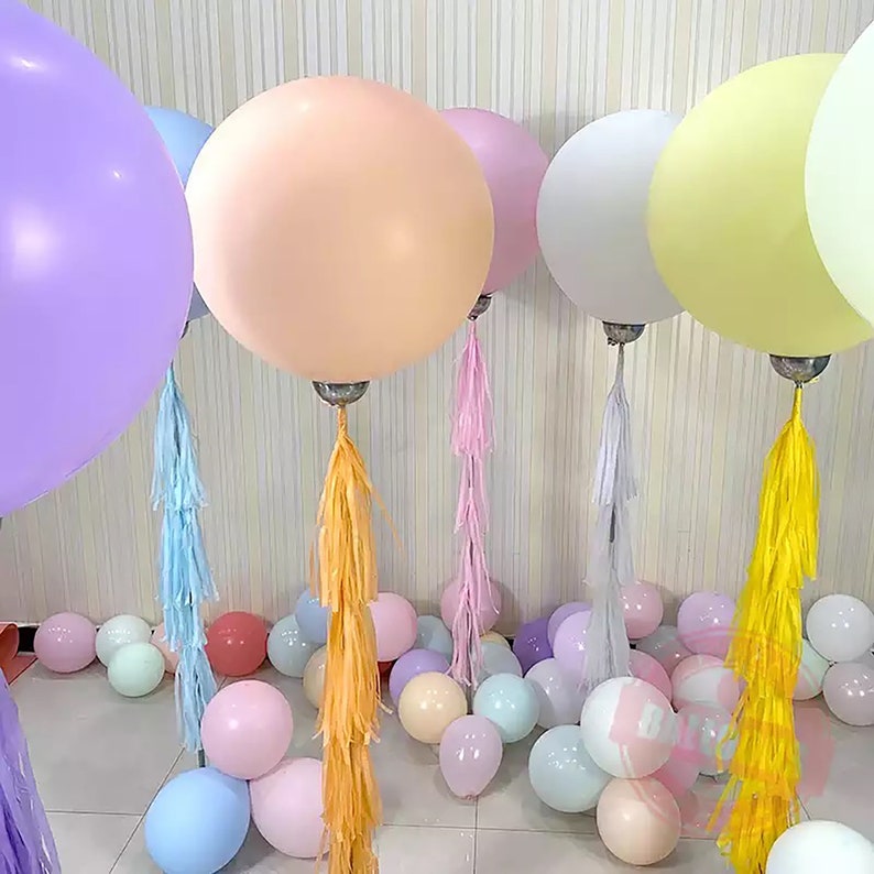 May include: A collection of large, round balloons in pastel colors, including peach, lavender, and yellow. Each balloon is adorned with a tassel and rests above smaller balloons scattered on the floor. Ideal for party decorations.