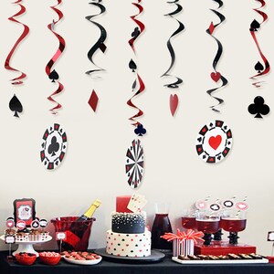 May include: Casino-themed party decorations. Red and black hanging swirls with card suit symbols and poker chip cutouts. A table is set with a cake, cupcakes, drinks, and other party supplies, all in a casino theme.