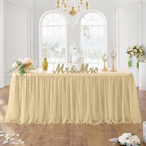 May include: A cream-colored table with a tulle skirt, decorated for a wedding. The table is set with a cake, champagne, and floral arrangements. Gold "Mr & Mrs" letters are displayed on the table. The room has large windows and a chandelier.