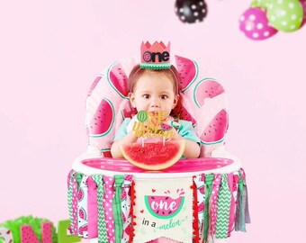 May include: A baby in a high chair decorated with a watermelon theme. The chair has a pink watermelon print, a banner that says "one in a melon", and a crown that says "one". The baby is holding a watermelon slice and a "Happy Birthday" cake topper.