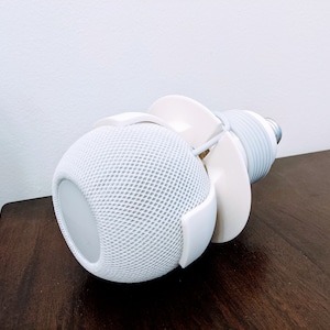 May include: White smart speaker with a white cord wrapped around it, mounted on a white stand. The speaker is on a dark brown wooden surface.