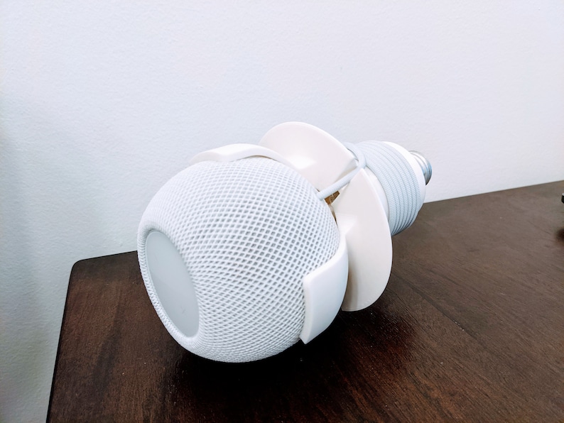 May include: White smart speaker with a white cord wrapped around it, mounted on a white stand. The speaker is on a dark brown wooden surface.