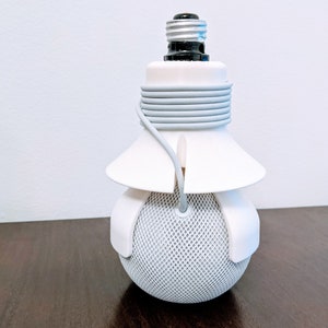May include: A white lightbulb socket adapter with a grey cord wrapped around it. The adapter is attached to a white speaker with a grey mesh covering.